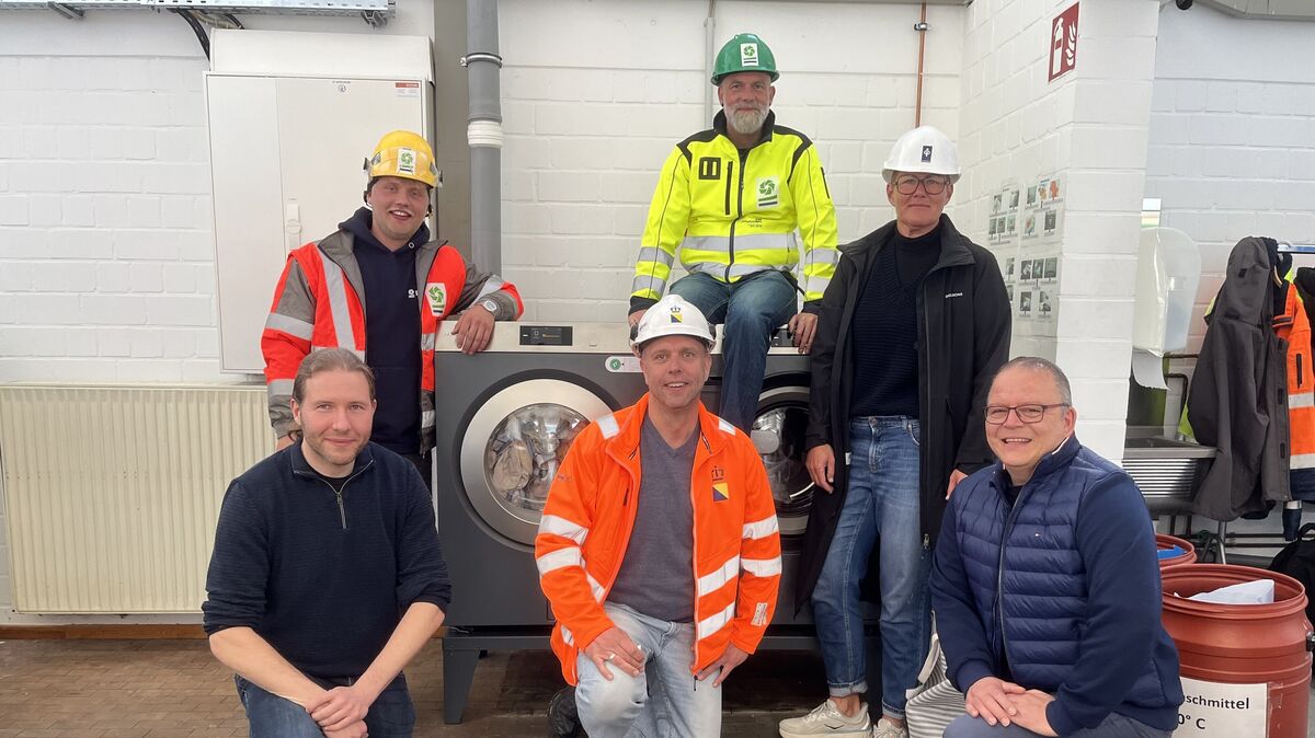Waschmaschine und Trockner laufen und sorgen dafür, dass täglich der Sand aus der Arbeitskleidung der Mitarbeiter auf der Großbaustelle gewaschen wird. Sehr erfreut über die Kooperation sind (v.l.) der stellvertretende Produktionsleiter der WWL, Patrick von Soosten, Thore Ruland (Nordsee-Nassbaggerei Deme), Rick Prummel (Boskalis Hirdes), Florian Schwarze (Nordsee Nassbaggerei Deme), Anja Albrecht (Depenbrock) und WWL-Produktionsleiter Alexander Twarok. Foto: Reese-Winne