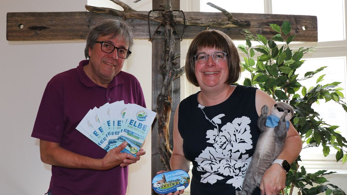 Kerstin Tiemann, Superintendentin des Kirchenkreises Cuxhaven-Hadeln, und Andreas Schoener, Referent für Öffentlichkeitsarbeit beim Kirchenkreis, vor dem Elbe-Wasser-Kreuz, das als ökumenisches Friedenssymbol regelmäßig von Ort zu Ort bei den Elbe-Kirchentagen weitergereicht wird. Zusammen mit Maskottchen Elbi weisen sie auf das umfangreiche Programm hin. Die Fischdose "Luther bei die Fische" dient als kulinarisches Werbemittel. Foto: Mangels